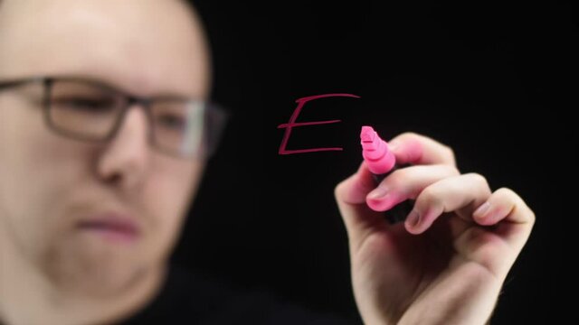Formula Equivalence of mass and energy on a transparent surface. The face and hand of a man with a marker on a black background. Red formula text on the board.