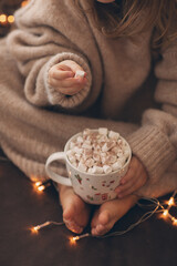 Child bare feet and hands hold cup hot cocoa marshmallow close up garland lights.flaffy fuzzy warm knitted beige sweater. Christmas concept, holiday.Happy New Year.Child girl sitting on sofa.Vertical