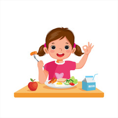 Cute little girl eating breakfast with bread, egg, broccoli and holding sausage with fork showing okay sign.