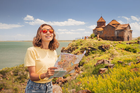 Young woman with map is looking for directions to the sights against the background of the famous Hayravank Monastery and Sevan Lake.