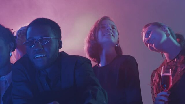Medium close up with slowmo of young Black man in trendy glasses rapping at camera during party with friends at night club with neon lighting and smoke