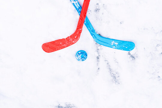 Children's Hockey Sticks Crossed On The Ice. Red And Blue Hockey Stick Crossed And Lying In The Snow. It Symbolizes The Opposing Team And Their Sports Fight, Passion And Desire To Win