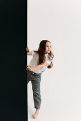 little girl 8 years old, in the studio on a white background, she looks out from behind a black wall that divides the white space