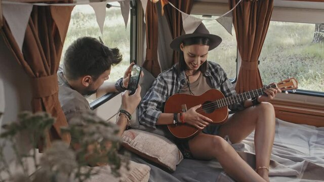 Medium shot slowmo of happy young woman sitting in camper and playing on ukulele while her boyfriend looking through open window and taking photo on mobile phone