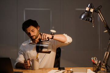 White bearded man drinking coffee while working with laptop in evening