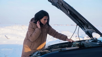 Upset young woman looks at dead engine standing near broken car with open bonnet on snowy road in...