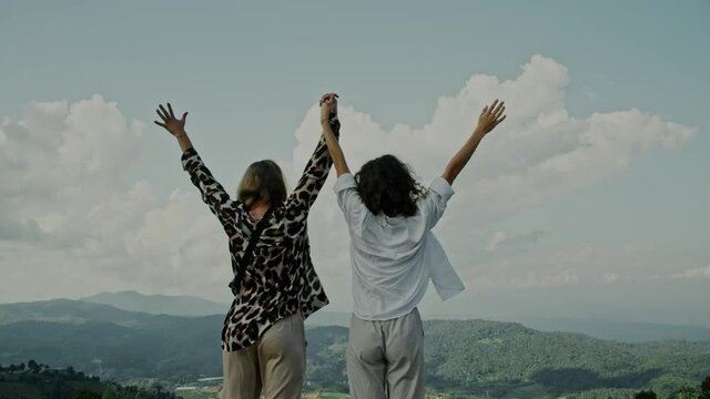 Slow motion, close-up of two young women standing on the edge of a cliff and raising their hands up against a mountain view. The happy girls are enjoying their success and the stunning view.