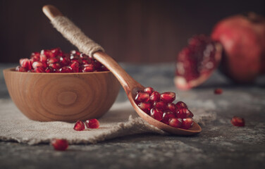 Pomegranate seeds lying in a wooden spoon, and in a wooden plate on a linen napkin. In the background are pomegranates with a slice of pomegranate.