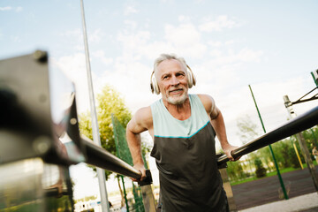 Grey senior sportsman listening music while working out on parallel bars