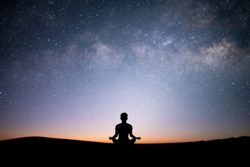 Silhouette of young female sitting practices yoga and meditating in lotus position alone on top of the mountain with beautiful night sky, star and Milky Way. She felt calm and happy.
