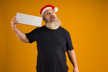 man in santa claus hat with a beard, black t-shirt, holding a white box on his shoulder and looking up against a yellow background, mockup