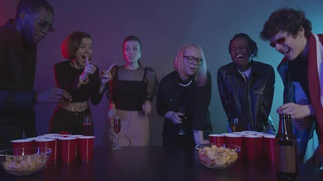 Slowmo shot of 6 joyful multiethnic friends divided in two teams having fun while playing beer pong at home party