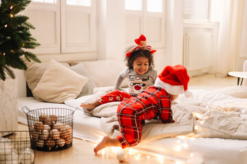 Multi-racial African American little curly-haired children in Christmas pajamas and Santa Claus hats are playing opening gifts in anticipation of the holiday in the New year vacations. Selective focus