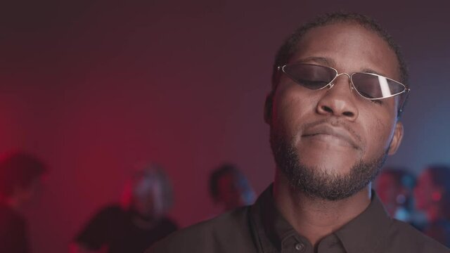 Chest up slowmo shot of relaxed African-American guy in trendy glasses dancing slowly with eyes closed at party in night club