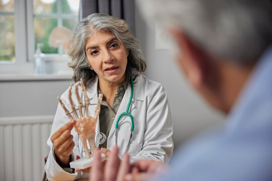 Mature Female Doctor Meeting With Male Patient Discussing Joint Pain In Hand Using Anatomical Model