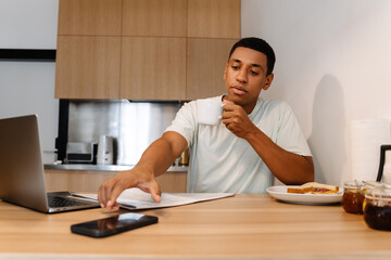 Black man drinking coffee while working with cellphone and laptop