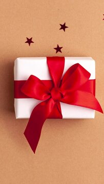 White gift box with a red satin ribbon bow tied on brown background with red stars shapes. Stop motion vertical animation Christmas Holidays and present concept flat lay
