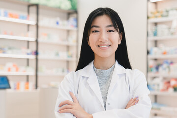 Cropped photo of asian female confident young pharmacist druggist in white medical coat looking at camera standing with arms crossed at the cash point desk in pharmacy drugstore