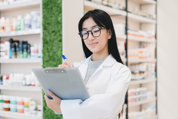 Asian female young pharmacist druggist in white medical coat writing on clipboard side effects, active substance, prescriptions standing at pharmacy drugstore