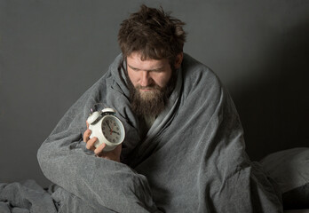 a sad shaggy young man is sitting covered with a blanket with an alarm clock in his hand. the...