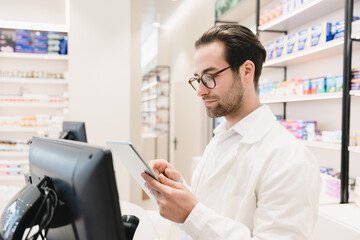 Obraz na płótnie Canvas Side view of caucasian male young pharmacist druggist checking drugs medicines pills remedies on prescription using digital tablet at cash point desk in drugstore pharmacy