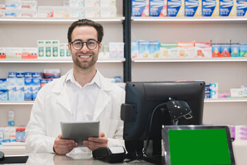 Confident smiling male caucasian druggist pharmacist using digital tablet standing at cash point...