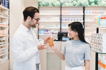 Consultation. Caucasian male young pharmacist druggist advising medicines, pills, drugs, painkillers, vitamins to a female asian customer client, buyer in drugstore