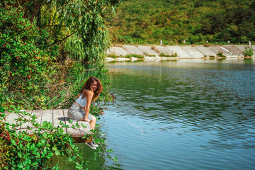 A charming young woman in a skirt is sitting on a wooden bridge against the background of nature with a lake in the forest. Travel, freedom, the concept of an active lifestyle.