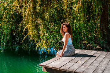 A charming young woman in a skirt is sitting on a wooden bridge against the background of nature with a lake in the forest. Travel, freedom, the concept of an active lifestyle.