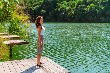 A woman stands on a wooden pier, relaxing enjoying the scenery of a forest and a mountain lake.