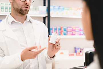 Male pharmacist druggist advising talking telling about new medicines pills remedies, side effects to a female customer patient holding a jar with mockup. Pharmacy consultation