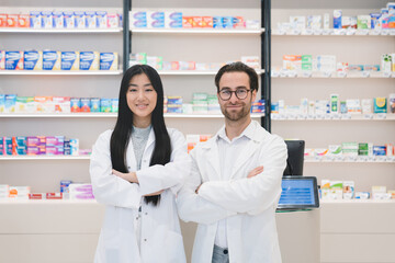 Two confident successful young pharmacists druggists in white medical coats standing with arms crossed at the cash point desk in pharmacy drugstore
