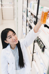 Female asian pharmacist druggist in medical white coat stretching to pills, vitamins, painkillers, medicines at pharmacy shelves in drugstore