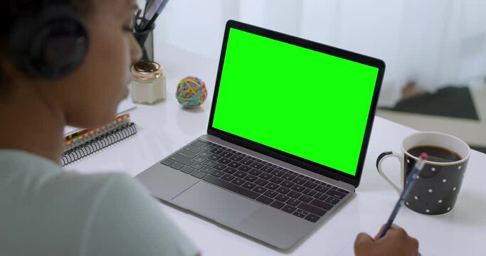 Over the Shoulder Shot of African American Woman Using Headphones Working or Studying on Laptop Computer with Green Screen. Chroma Key Online Course, Remote Learning.