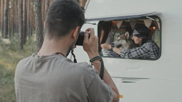Slowmo medium shot of young man with digital camera taking photos of woman with husky looking out open window of camper parked in forest