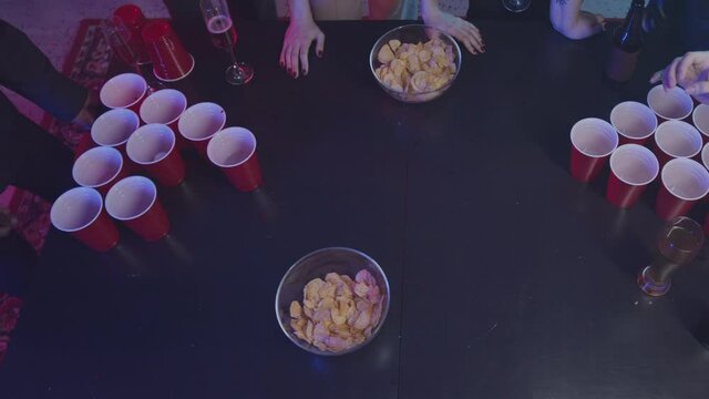 Top-view slowmo shot of table with red cups and chips in neon lights. Friends playing beer pong at home party