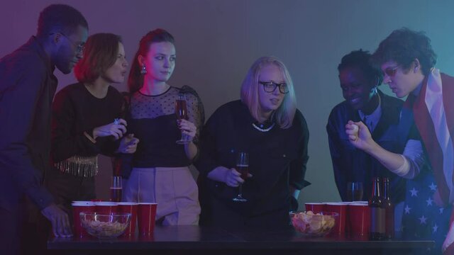 Slowmo shot of diverse young people divided in two teams playing beer pong and having fun together during home party in room with neon lights