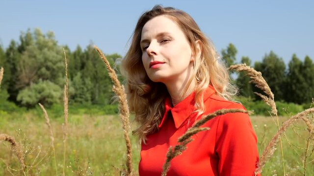 View through the dry grass to a portrait of a blonde woman in a red dress in a field. Slow motion 4K