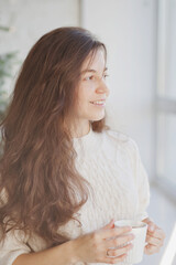 Beautiful young woman drinking coffee or tea near christmas tree in the living room at home closeup