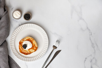 Black Caviar on pancakes with sour cream Holiday Appetizer on white plate and marble background...