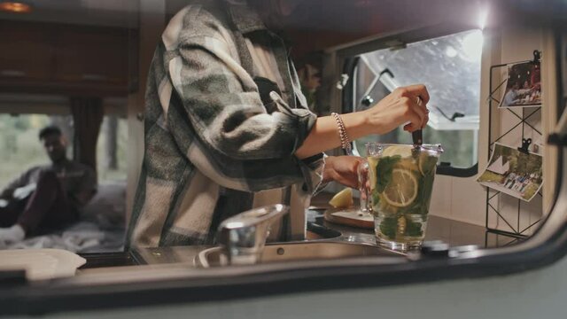 Tracking slowmo mid-section shot of young woman making refreshing lemon and mint water in small kitchen in camper on sunny summer day. Man playing guitar in background