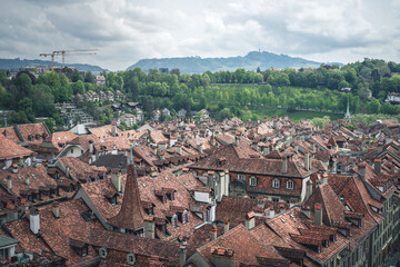A view of the cityscape of Bern, Switzerland with stone rooftops, streets below, and the farmer's market - a cloudy spring day with green trees and hills in the background