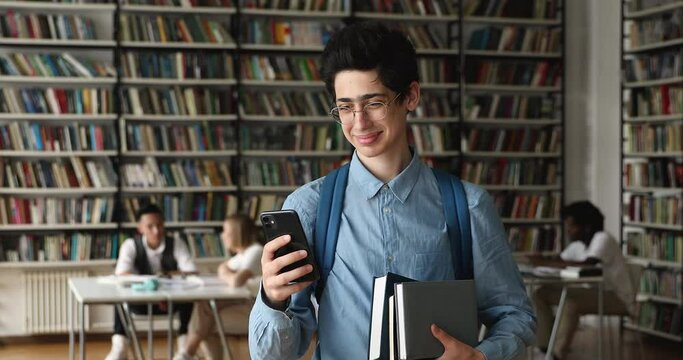 Highschool guy student in eyeglasses holds textbook and cellphone check messages, chatting while standing in university library. Gen Z, usage of new mobile application, online class timetable concept