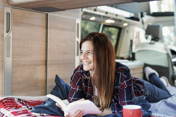 Mature caucasian woman reading book and drinking tea inside mini van camper during road trip vacation