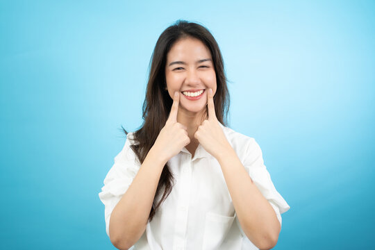 Front view half-body image of a happy Asian woman standing using an index fingers touching the corners of mouth while smiling broadly showing her beautiful white teeth isolated on turquoise background