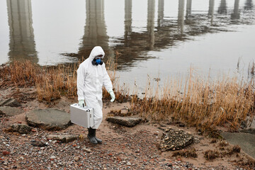 Obraz premium Full length portrait of worker wearing hazmat suit walking by water outdoors carrying samples case, industrial waste concept, copy space