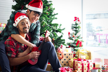Adorable young LGBT couple sharing special moment together on Christmas holiday, Asian gay male lover sitting in living room, relaxing and singing a song while playing ukulele to celebrate Christmas