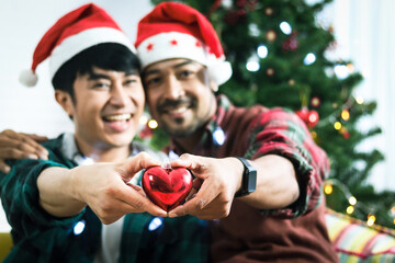 Adorable smiling young LGBT couple holding red heart and giving it to the camera, Asian gay male lover celebrating and sharing special moment together on Christmas holiday in living room
