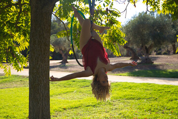 Blonde woman and young gymnast acrobat athlete performing aerial exercise on air ring outdoors in park. Flexible woman in red costume performs poses of circus performers dancing with hips.