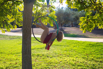 Blonde woman and young gymnast acrobat athlete performing aerial exercise on air ring outdoors in park. Flexible woman in red costume performs poses of circus performers dancing with hips.